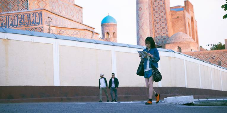 Eine junge Frau geht mit einer Zeitung in der Hand eine Straße entlang. Sie trägt auffallend gelbe Schuhe und eine Jeansjacke. Im Hintergrund sind orentalisch angehauchte Gebäude zusehen, deren Wände mit Mosaiken und Fresken übersäht sind.